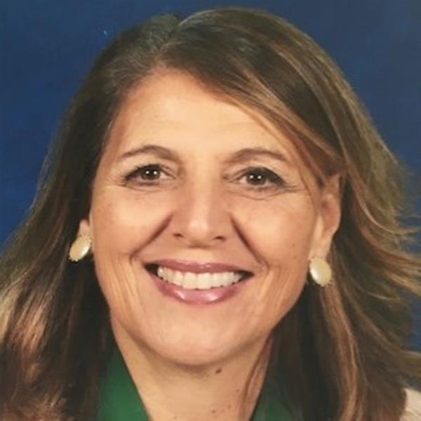 Portrait of former principal Mary Ellen Sanchez smiling for a headshot photo, seen with hair down, wearing green shirt and large, round earrings.