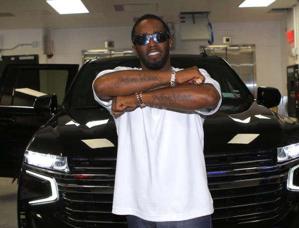 Rapper Diddy smiles and shows off New York tattoos wearing shades and a white tee, standing in front of an SUV.