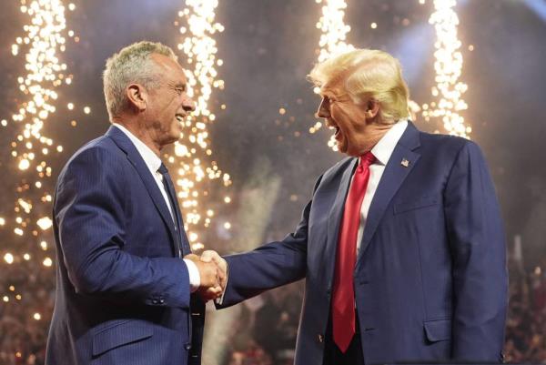 Former President Do<em></em>nald Trump shaking hands with Robert F. Kennedy Jr. on a brightly lit stage at a rally, with fireworks in the background.