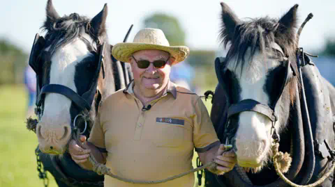 PA Gerry North from Offaly with horses Roo<em></em>ney (left) and Sunny at the Natio<em></em>nal Ploughing Championships. He is wearing a dark yellow polo shirt and a straw hat and sunglasses. He is holding each horse by the bridle.