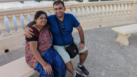 Atul Subhash with his mother in happier times - they are both sitting on a cream coloured bench outdoors. He is wearing a blue t-shirt, shorts, has what looks to be a camera bag around his neck, and is smiling at the camera with his arm around his mother. His mother is wearing a red traditio<em></em>nal dress and is also smiling at the camera</p>

<p>　　